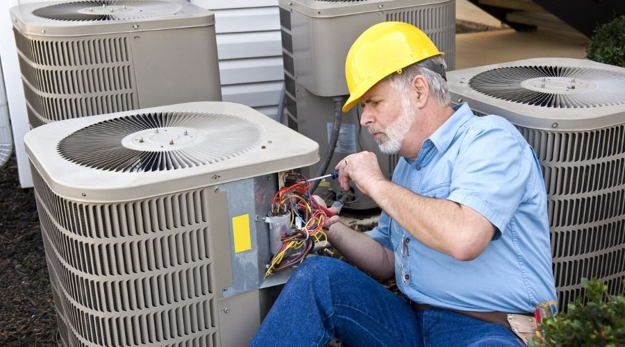 Technician in a yellow hard hat repairing air conditioning unit outdoors, focusing on electrical connections.