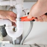 Plumber using adjustable wrench to repair plumbing pipes under a sink, emphasizing maintenance to prevent costly replacements.